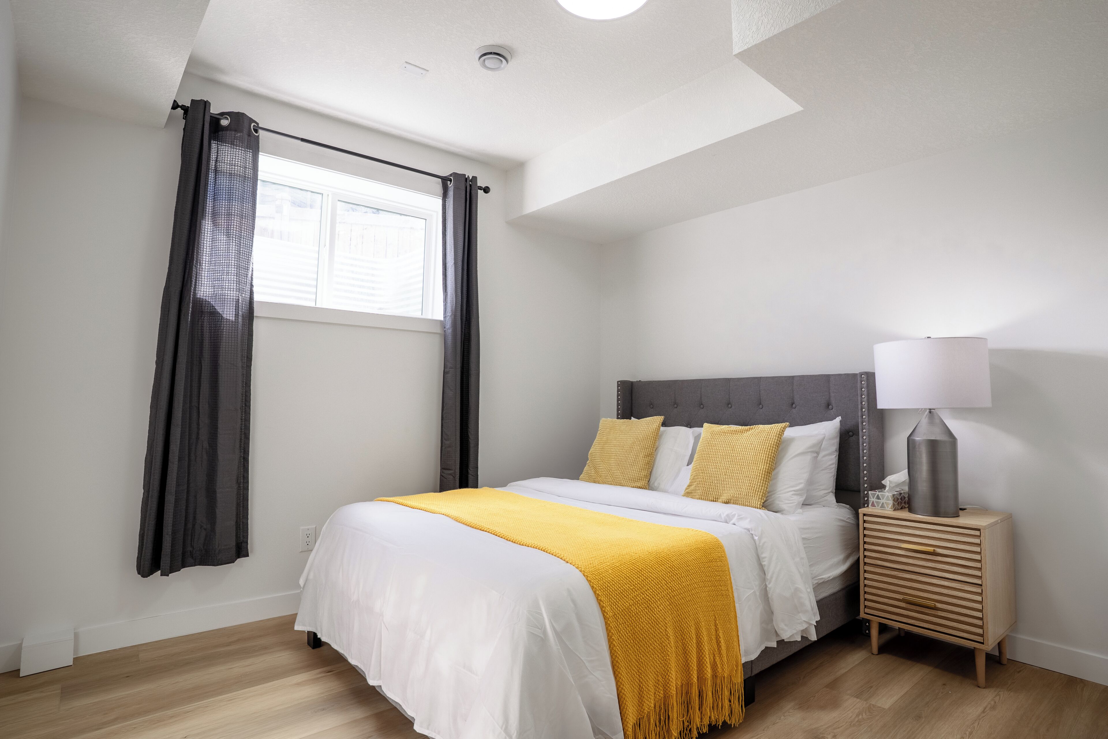 Um quarto com uma cama, uma mesa de cabeceira com luminária, uma janela com cortinas e uma luminária no teto.