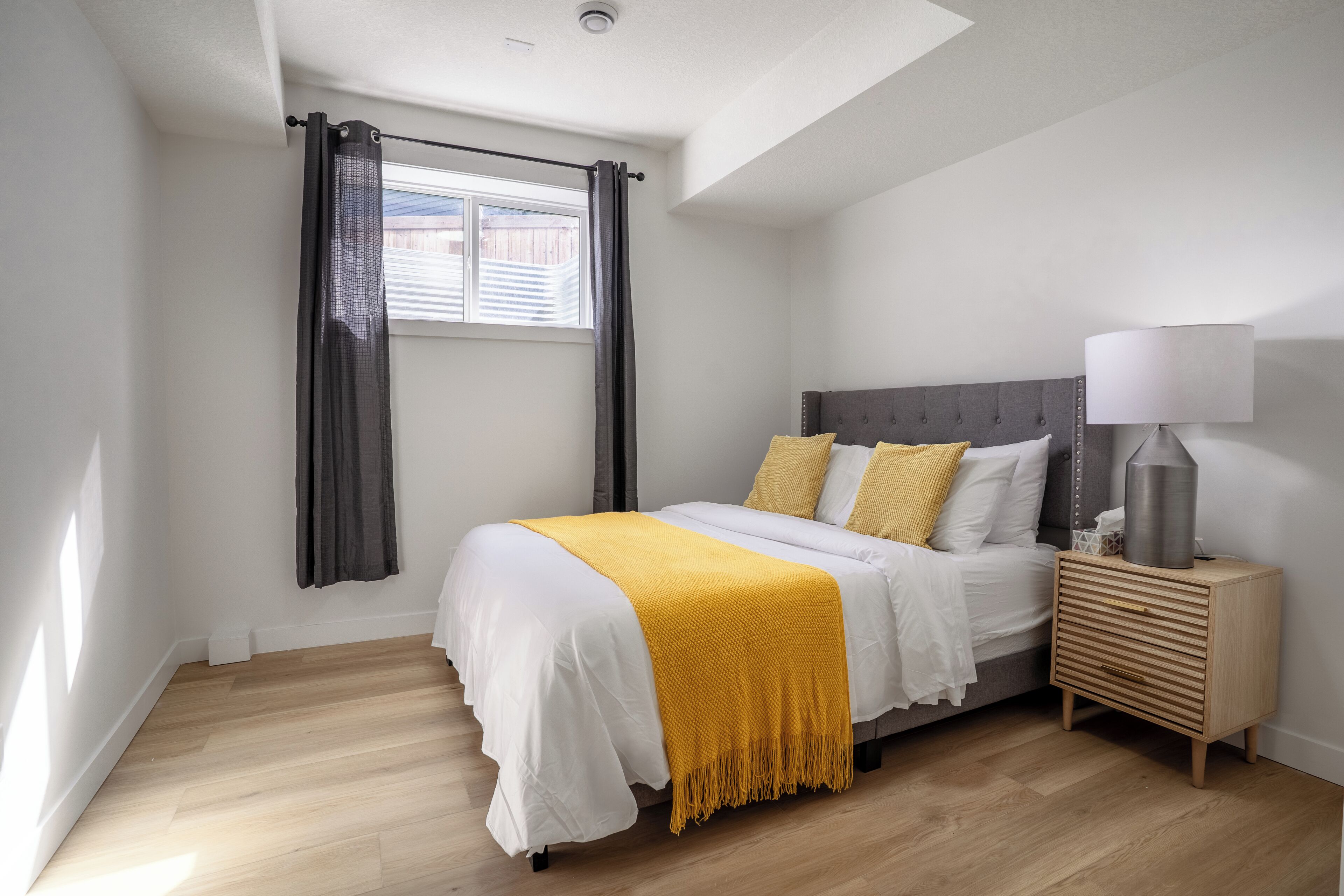 A bedroom with a bed, bedside table, lamp, and window with curtains.