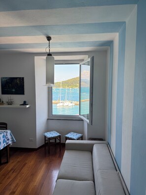 Ein Zimmer mit großem Fenster, einer Couch, einem kleinen Tisch und einem Wandregal.