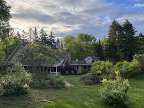 A two-story house with a stone chimney, surrounded by a lush garden and tall trees. - Maine Island Charm, 2 Miles to Hadley Pt Beach, Cafe, Coffee Shop & Winery. (Bar Harbor)