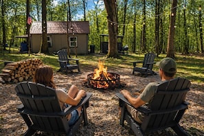 Duas pessoas sentadas perto de uma fogueira com cadeiras Adirondack, uma cabana pequena e uma área arborizada ao fundo.