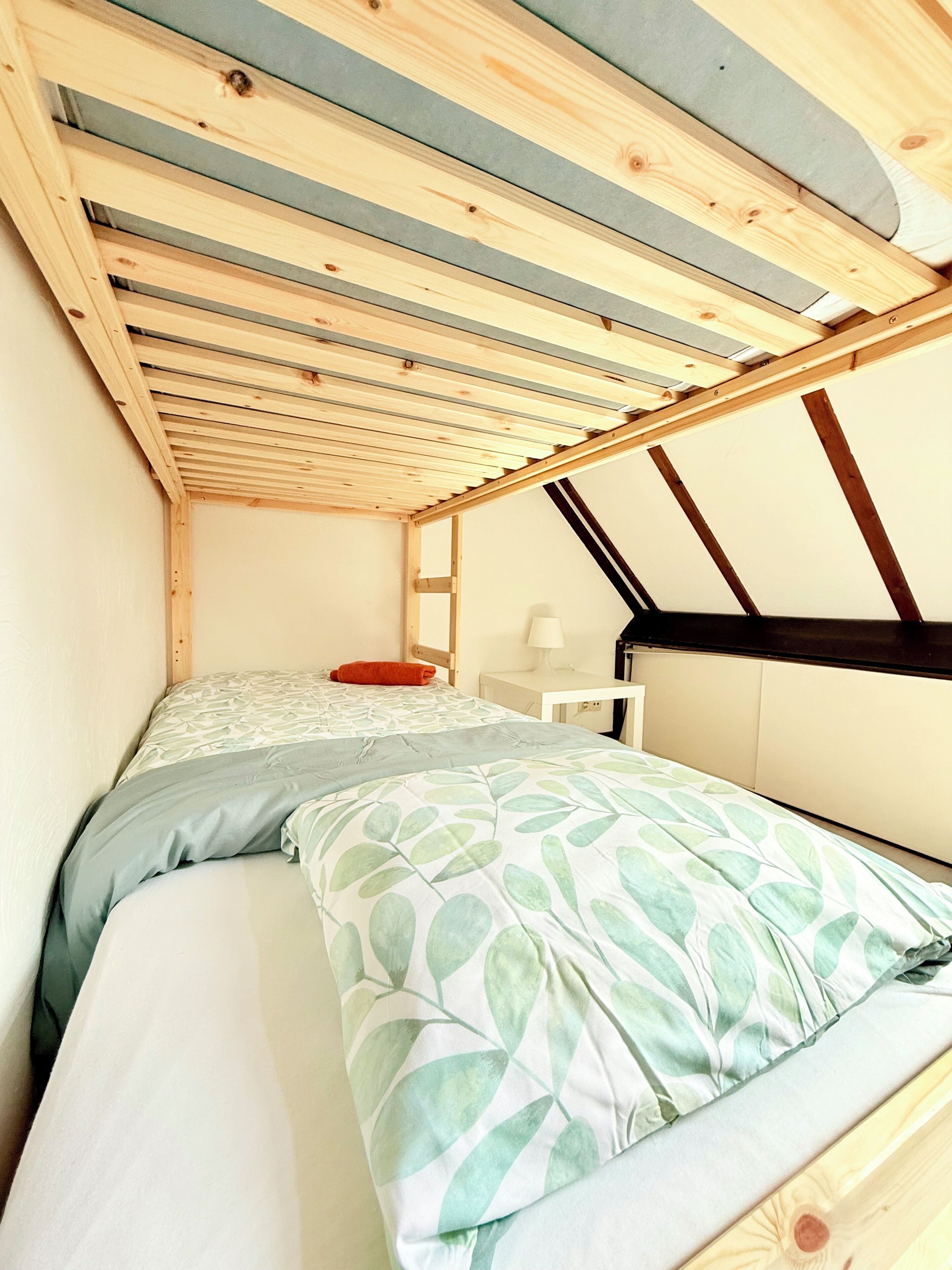 A small, single-bed room with a sloped ceiling, a bedside table, a lamp, and a window.