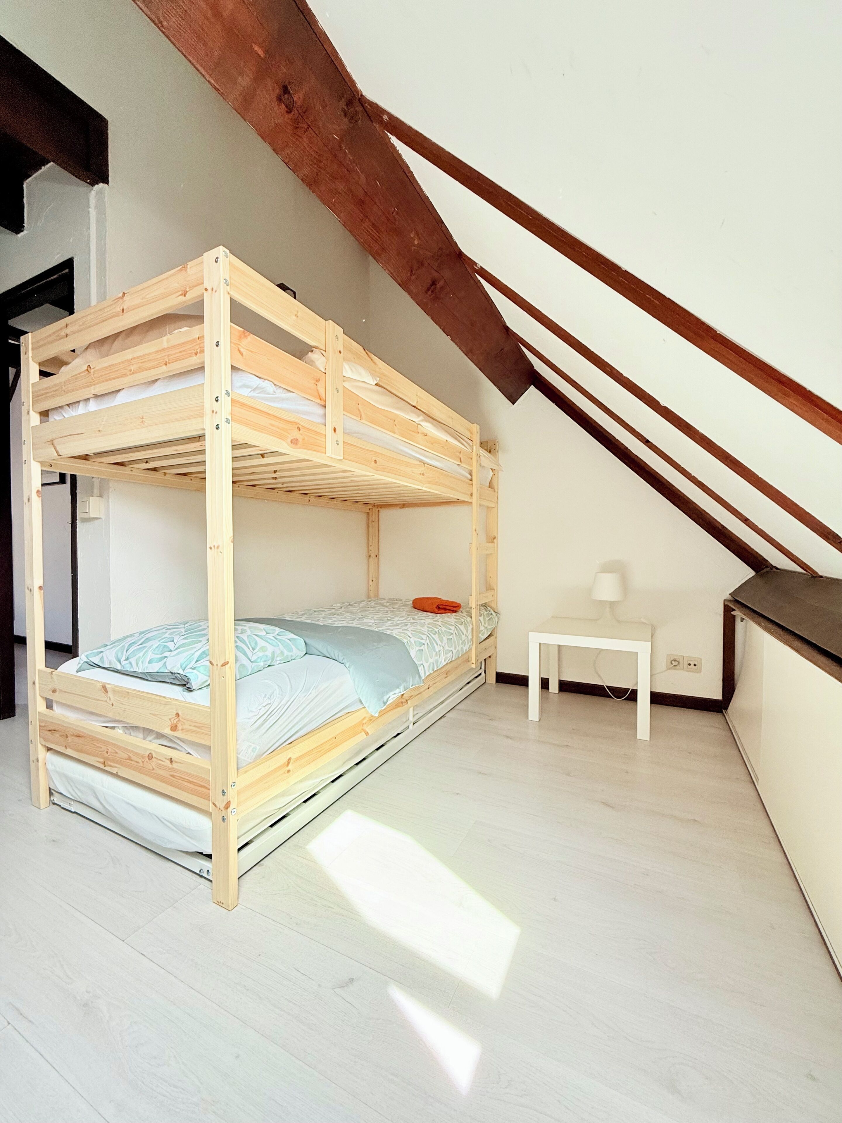 Un lit superposé dans une pièce dont le plafond est en pente, avec une petite table blanche et une lampe.