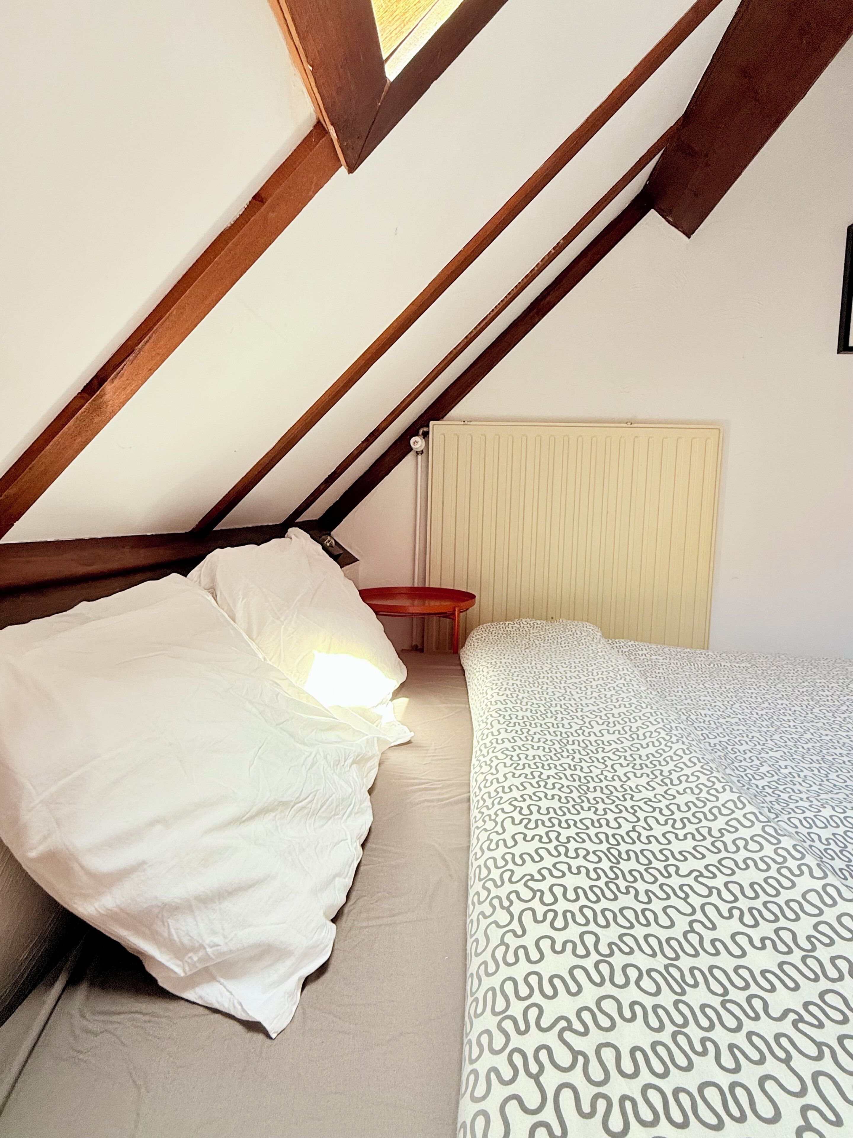 A bedroom with a sloped ceiling, a bed with white bedding, a patterned blanket, and a radiator.