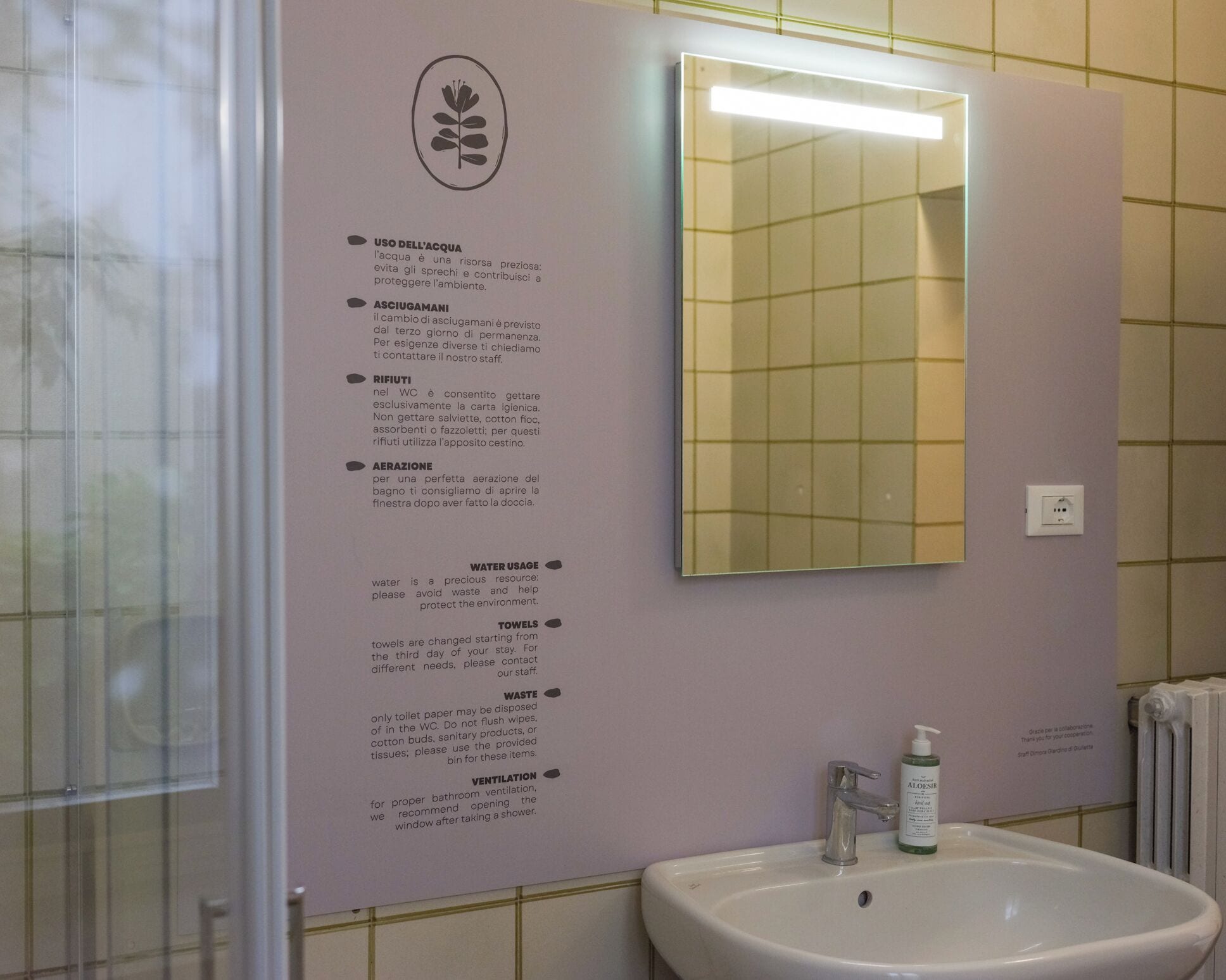 Un baño con lavamanos, espejo y carteles informativos sobre el uso del agua y las políticas de toallas.
