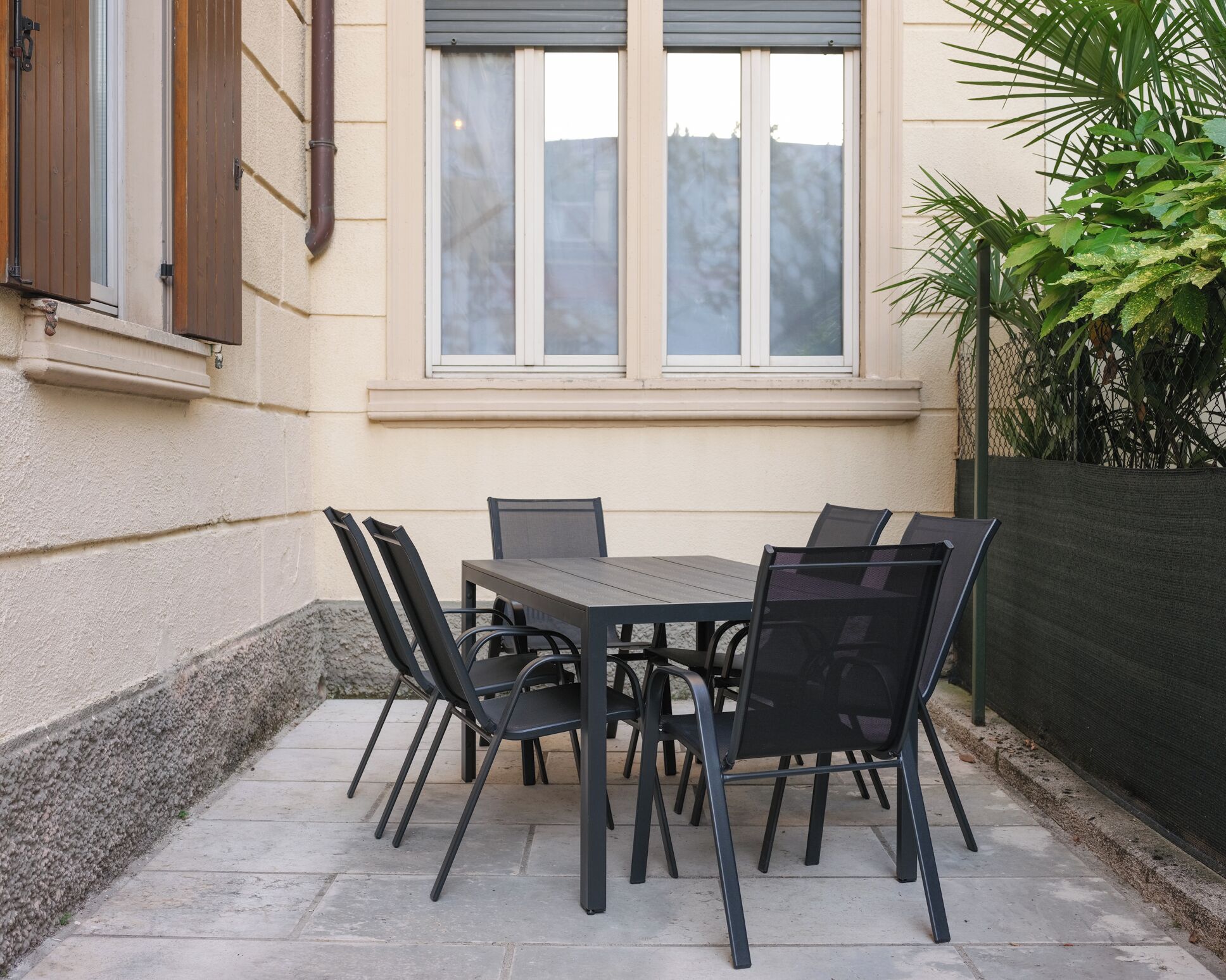 Un patio al aire libre con una mesa, sillas, una ventana con persianas y una planta.