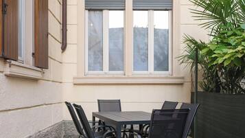 Un patio al aire libre con una mesa, sillas, una ventana con persianas y una planta.