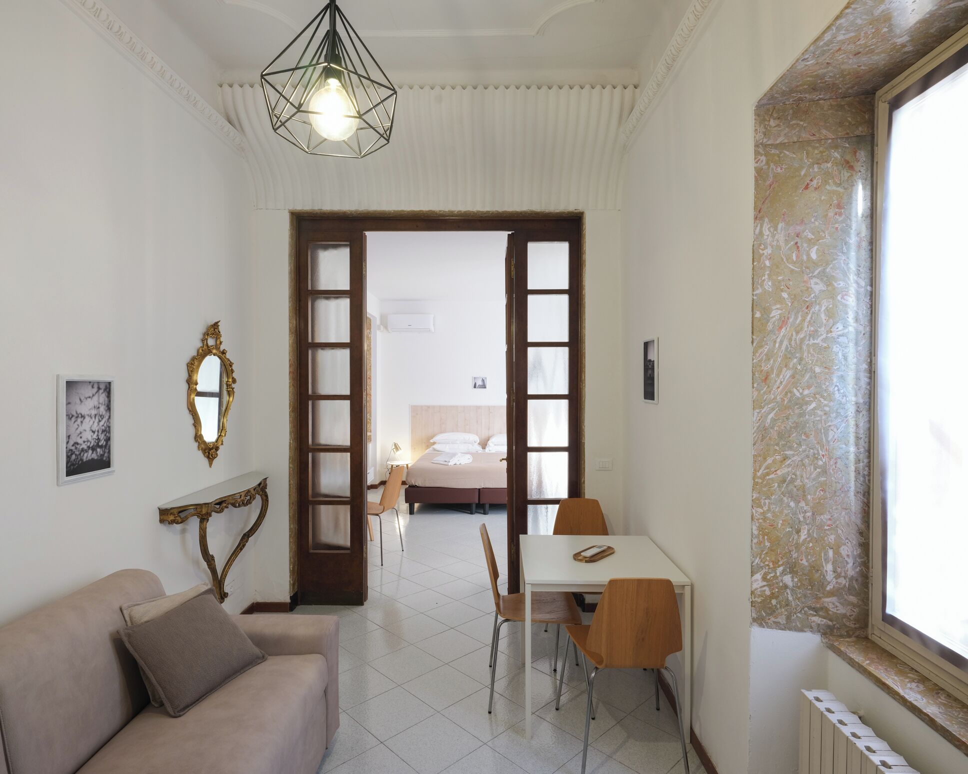 A hotel room with a bed, a dining table with chairs, a sofa, a mirror, and a hanging light fixture.