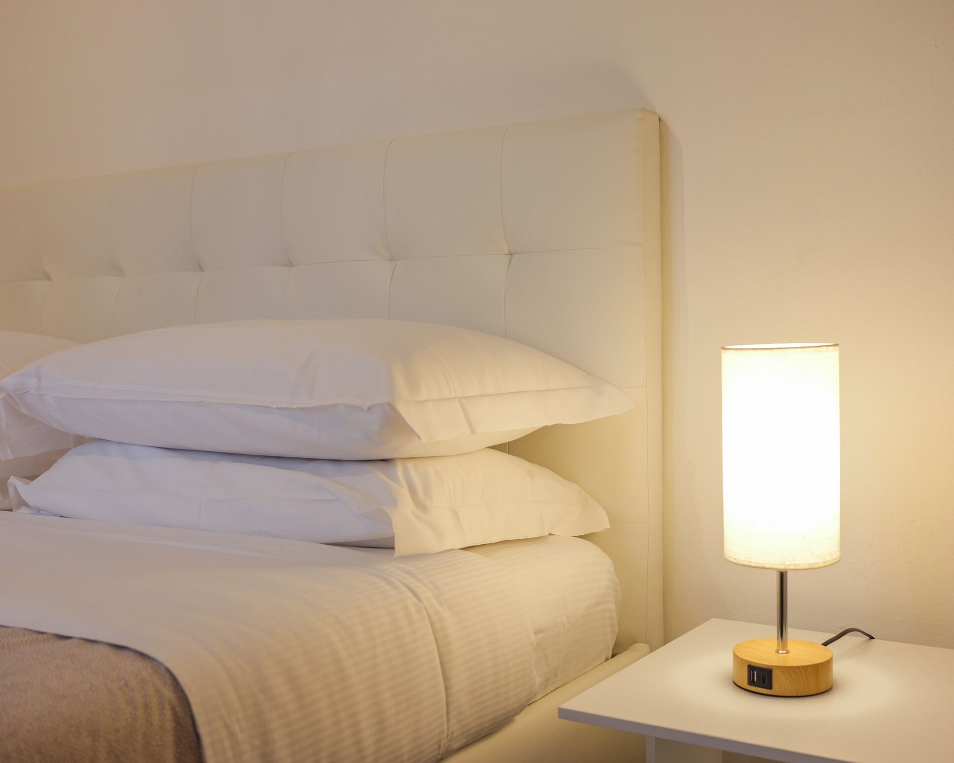 Un lit bien fait, avec du linge de lit blanc et des oreillers, une table de chevet avec une lampe, et une chambre au design épuré et simple.