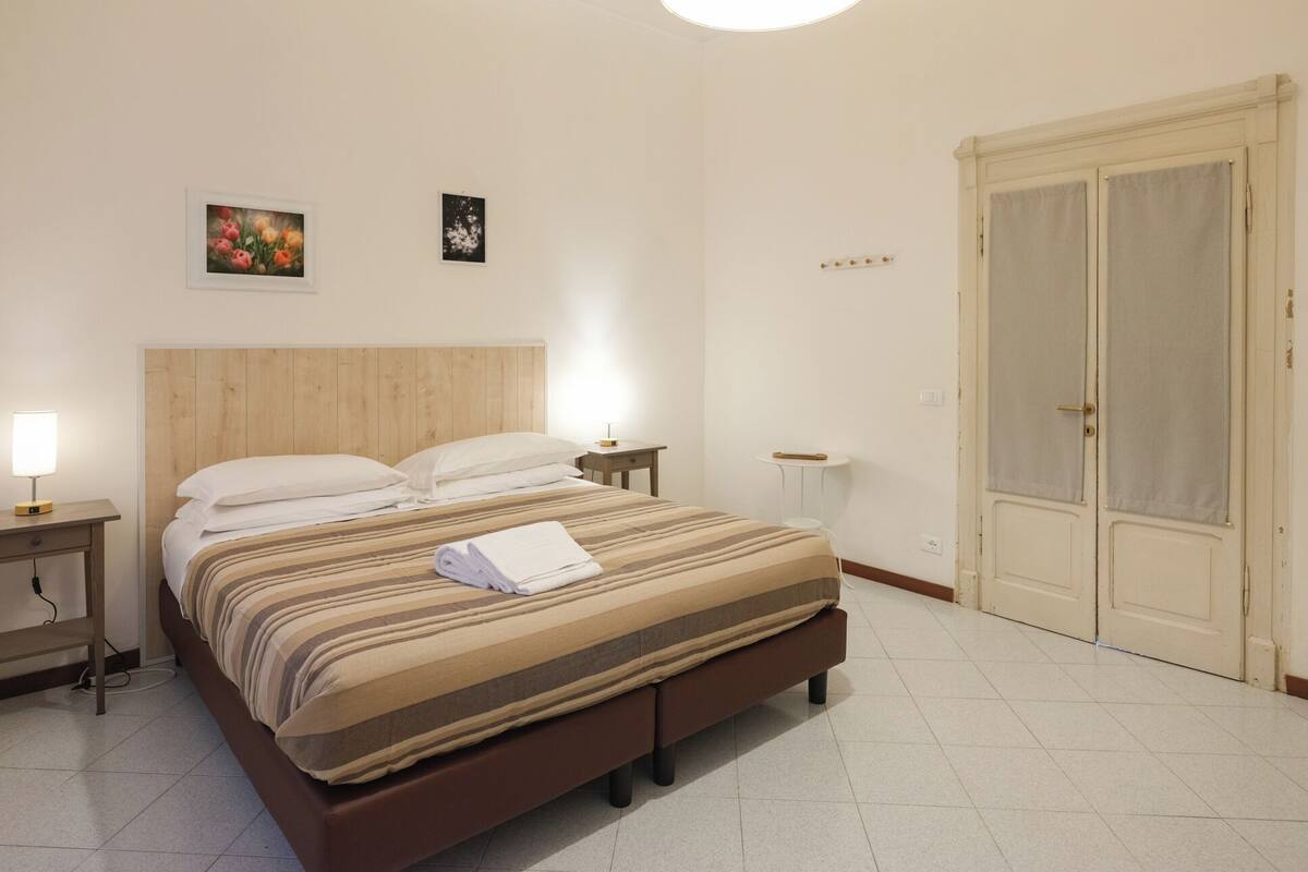 Un dormitorio con cama, mesitas de noche, lámpara, puerta y un aplique de pared.