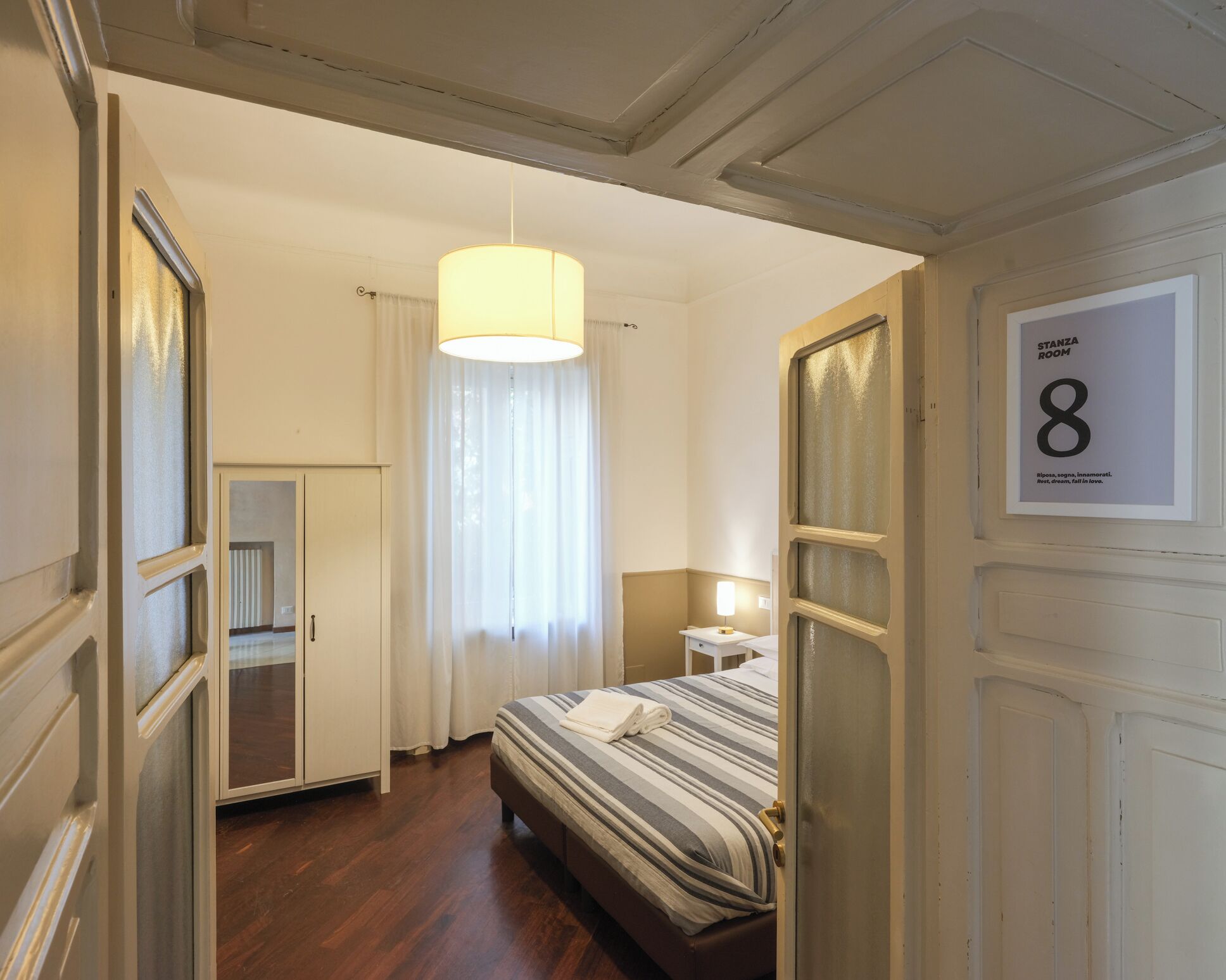 Ein Hotelzimmer mit Bett, Nachttisch, Lampe, einem Fenster mit Vorhängen und einer Tür mit Schild, auf dem Zimmer Nummer 8 steht.