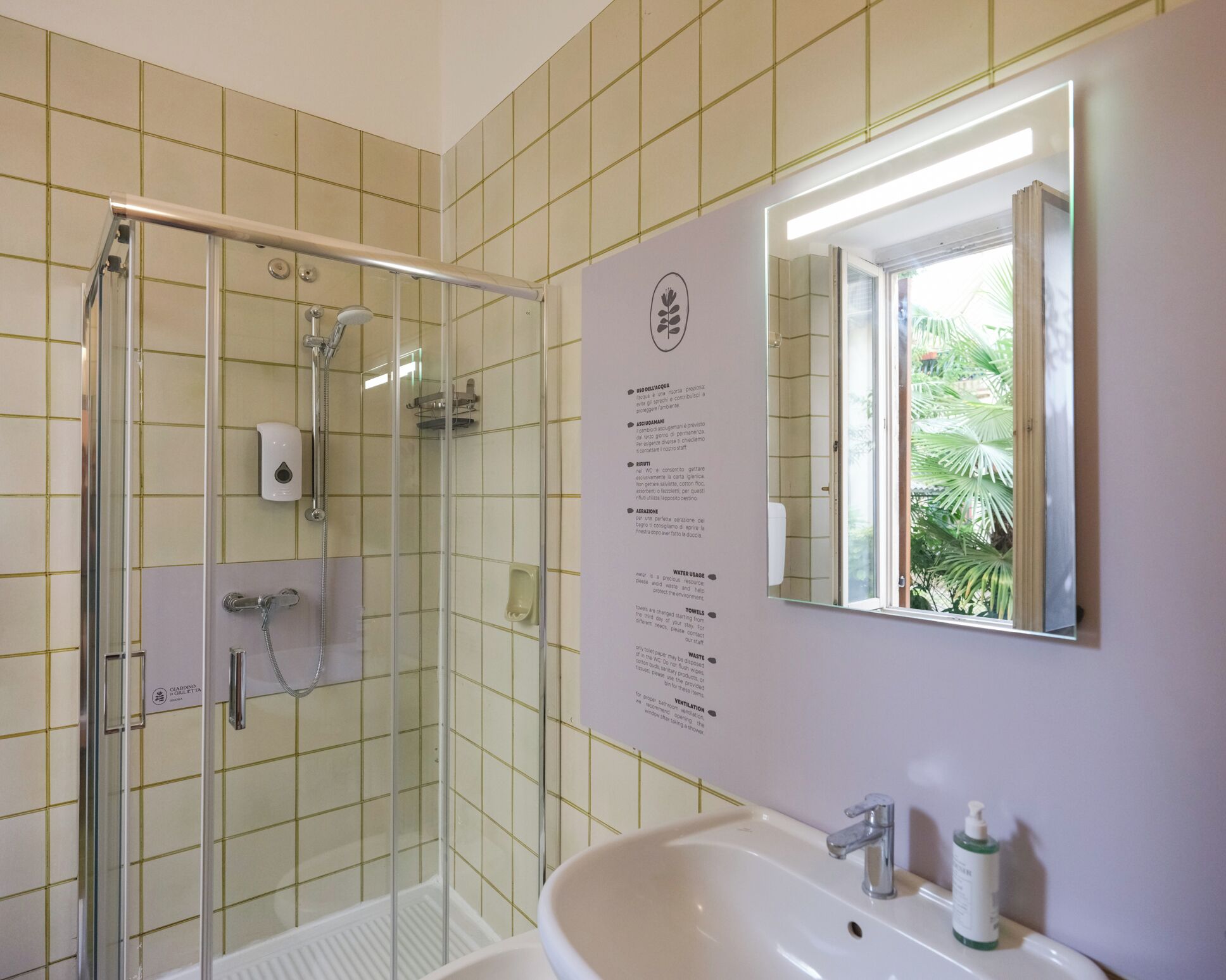 Un baño con ducha de vidrio, un lavamanos con espejo y un panel informativo en la pared.