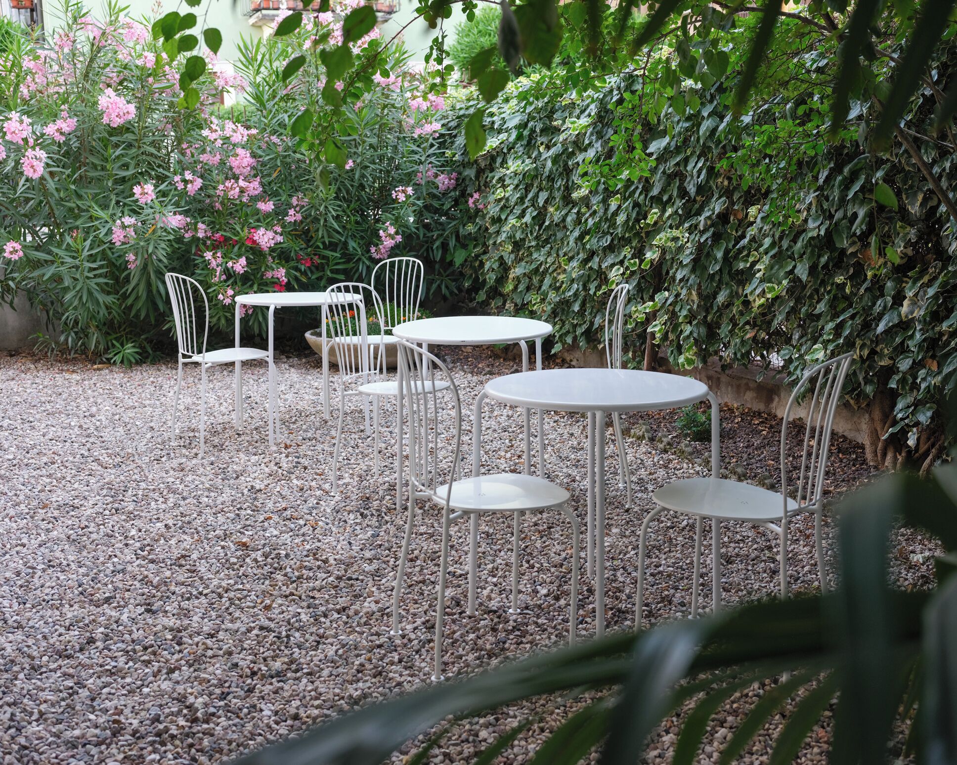 Un patio jardín con mesas y sillas de metal blanco, rodeado de grava y vegetación.