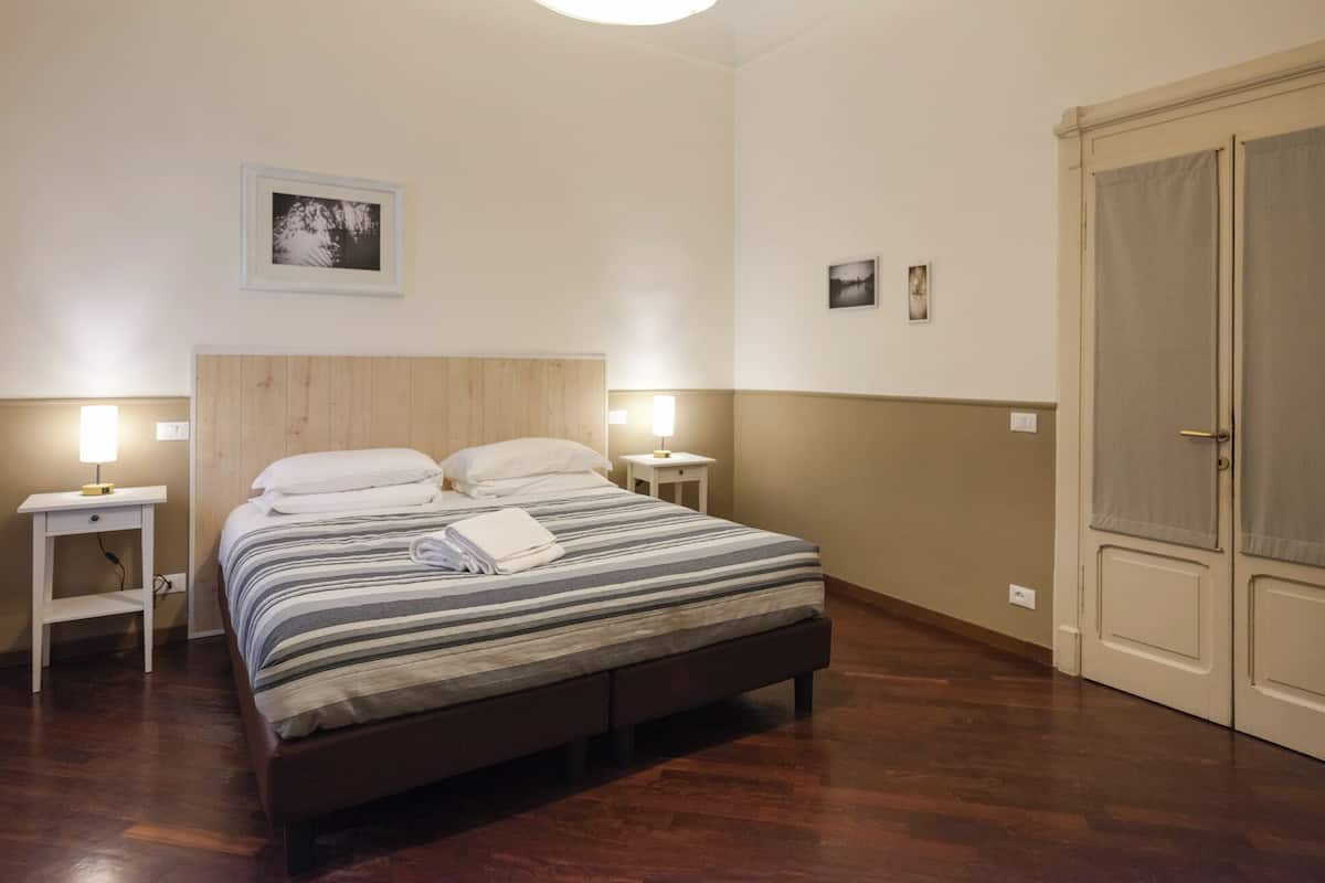 Habitación de hotel con cama, lámparas de noche, cabecera de madera, una puerta y un cuadro colgado en la pared.