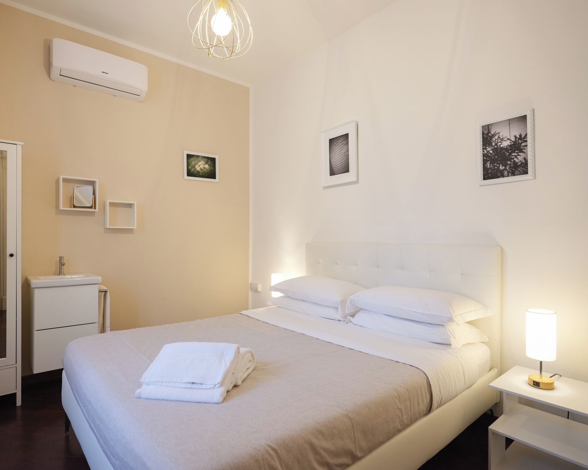 Quarto de hotel com cama, mesa de cabeceira com abajur, criado-mudo e ar-condicionado fixado na parede, além de quadros na parede.