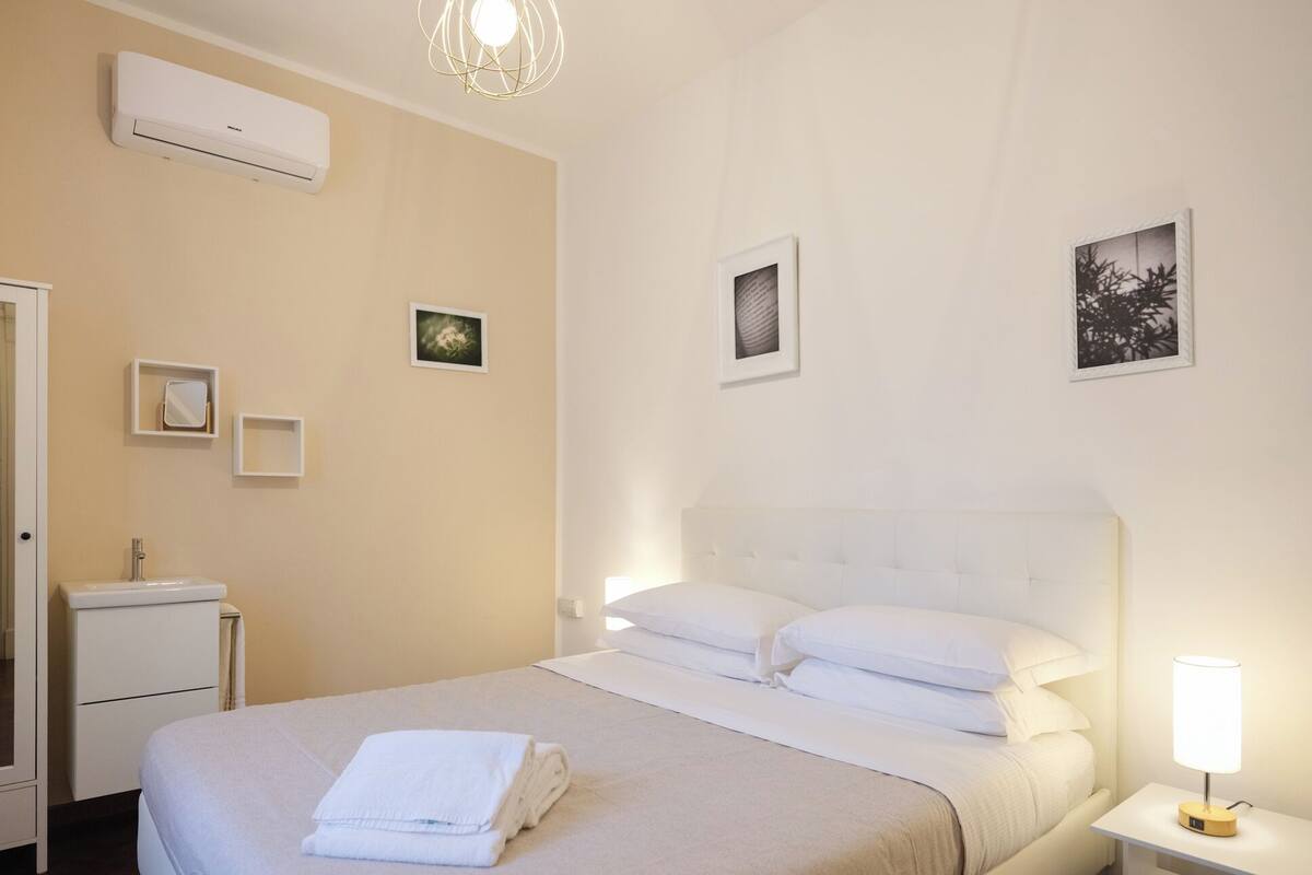 Habitación de hotel con cama, mesita de noche con lámpara, aire acondicionado de pared y cuadros enmarcados.