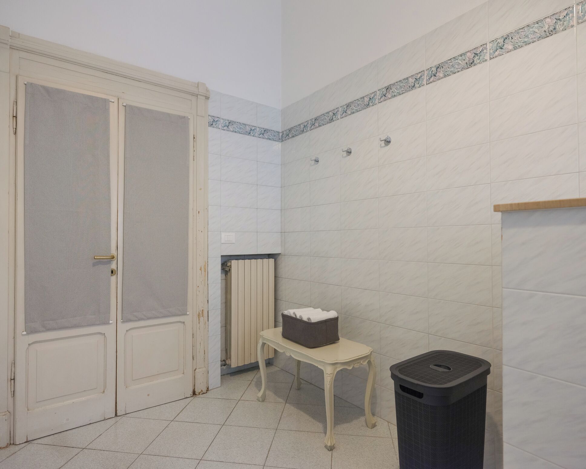 A bathroom with white tiled walls, a radiator, a small table with a basket, and a trash bin.