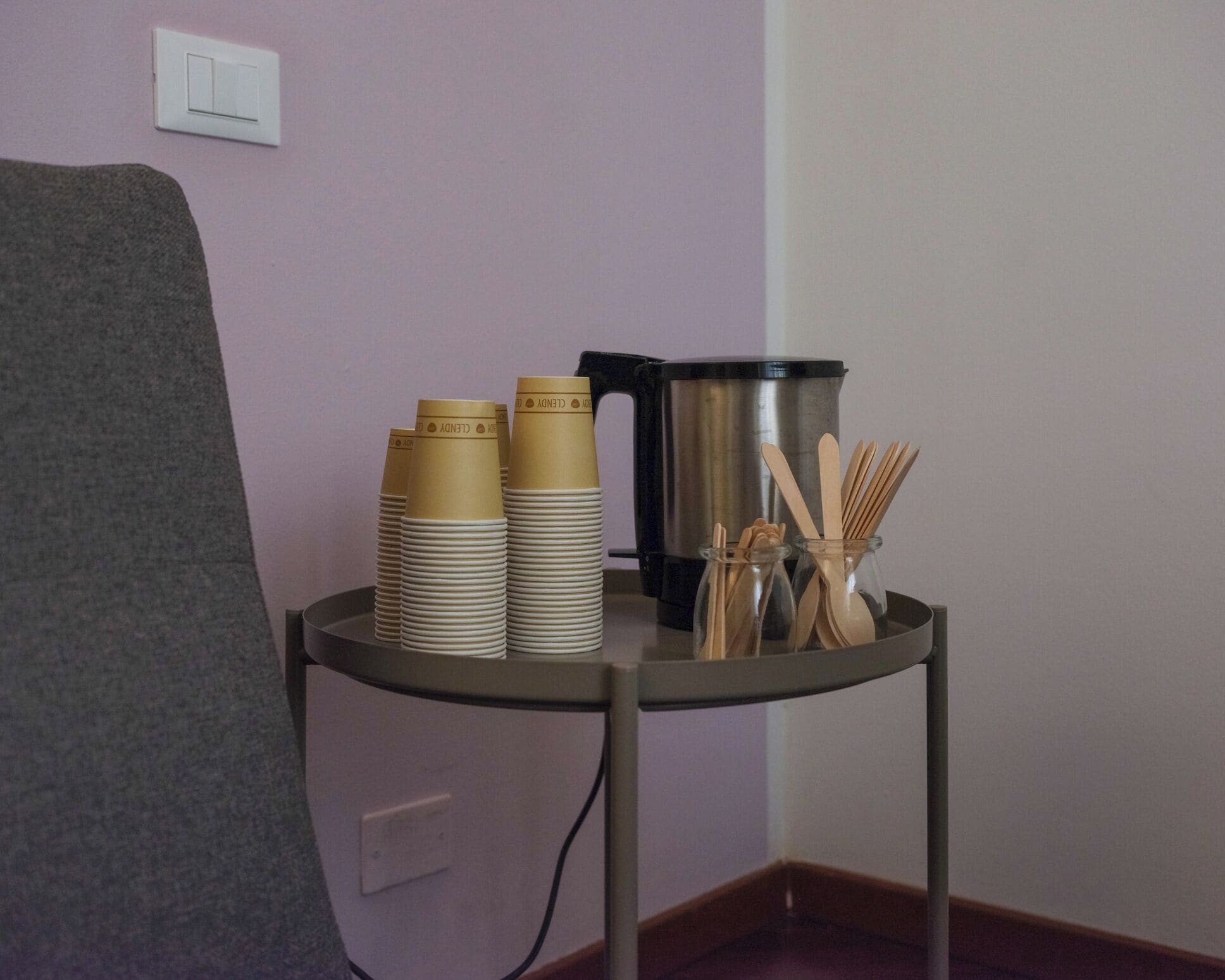 Un coin de pièce avec une chaise grise, une petite table sur laquelle sont empilées des gobelets en papier, un récipient noir et un bocal en verre contenant des ustensiles en bois.
