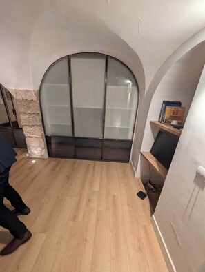 Ein Zimmer mit gewölbter Decke, einer Wand aus Stein, Holzboden, einer Schiebetür aus Glas und einem kleinen Bücherregal mit Büchern.