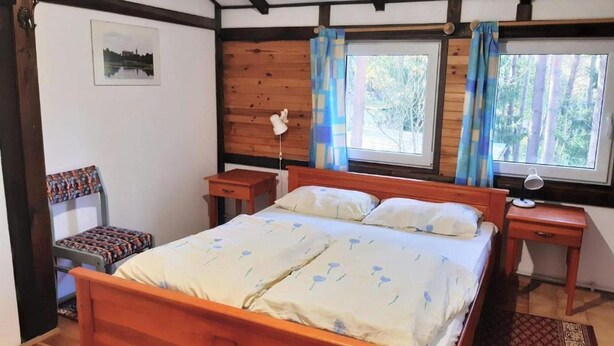 Un dormitorio con una cama de madera, dos mesitas de noche, una silla, una ventana con cortinas e imagen enmarcada en la pared.