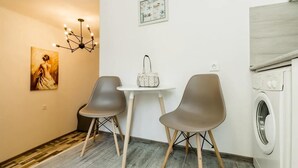 Una habitación pequeña con lavadora, una mesa y dos sillas, un candelabro y un cuadro en la pared.