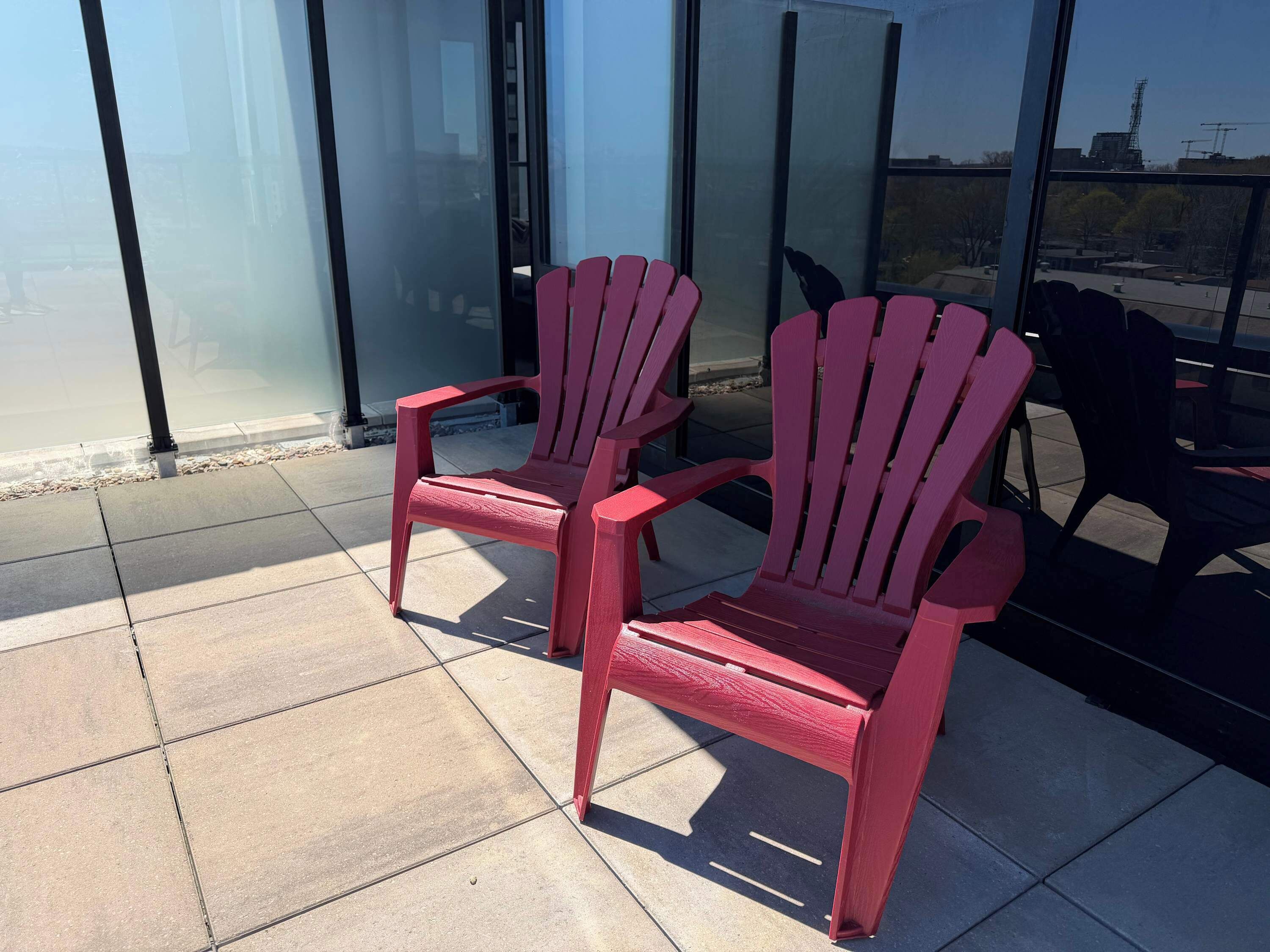 Two red Adirondack chairs on a tiled patio with a glass wall and a view of a building.