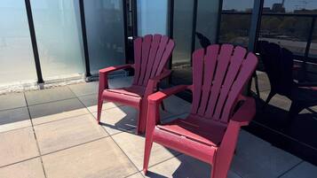 Two red Adirondack chairs on a tiled patio with a glass wall and a view of a building.