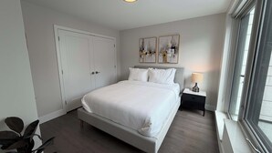 Een moderne slaapkamer met een groot bed, een nachtkastje met een lamp, een kledingkast en abstracte wanddecoratie.