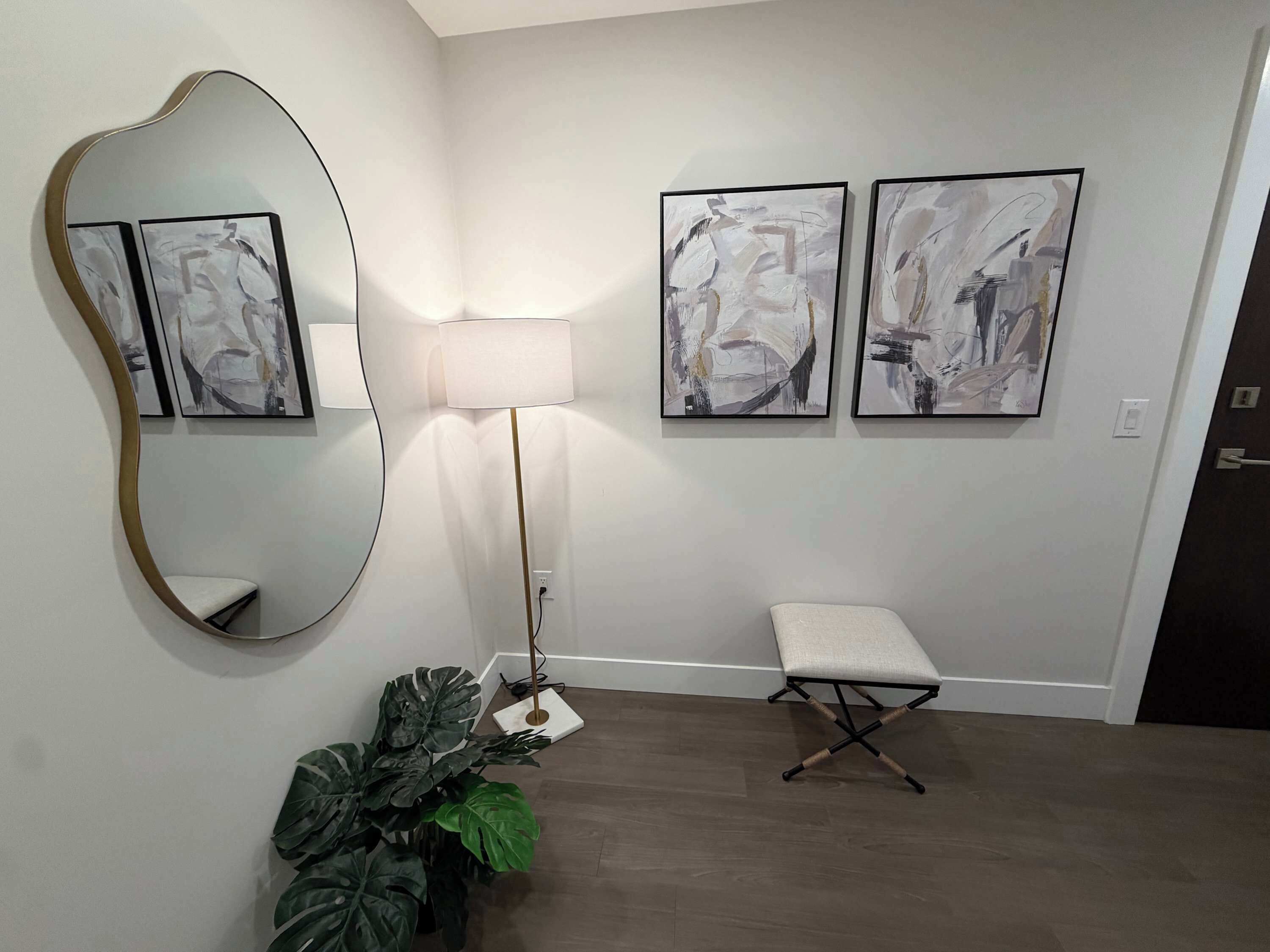 Una habitación con un espejo grande, arte abstracto en la pared, una lámpara de pie, un banco y una planta en maceta.