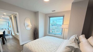 A bedroom with a large bed, a window with a city view, a bedside lamp, and a mirror on the wall. - Downtown Waterview, 2 BR/2 Bath with 6 appliances (Halifax)