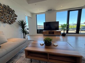 Un soggiorno moderno con divano, tavolino in legno, televisore piatto su supporto in legno e ampie finestre con vista su edifici e vegetazione.