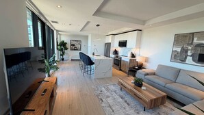 A modern living room with a TV, sofa, coffee table, and a kitchen area in the background. - Northend HFX 2 BR/ 2 Bath Penthouse with waterview (Halifax)