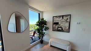 A modern interior with a large window, a round mirror, a potted plant, a bench, and a framed abstract painting. - Northend HFX 2 BR/ 2 Bath Penthouse with waterview (Halifax)
