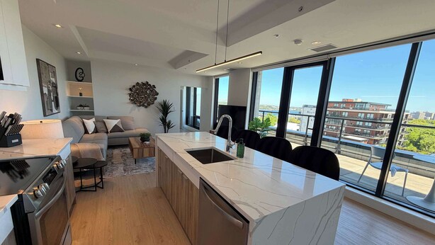 A modern kitchen with a large island, a dining area with a sofa, and large windows offering a city view. - Northend HFX 2 BR/ 2 Bath Penthouse with waterview (Halifax)