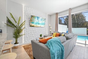 Ein modernes Wohnzimmer mit grauem Sofa, einem großen Gemälde von einem Strand, einem Essbereich mit Marmortisch und Blick auf einen Swimmingpool.