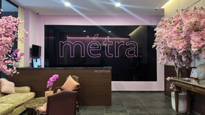 A reception area with a reception desk labeled 'RECEPTION,' a sofa, a chair, and a vase with flowers. - Metra Tower (Phnom Penh)