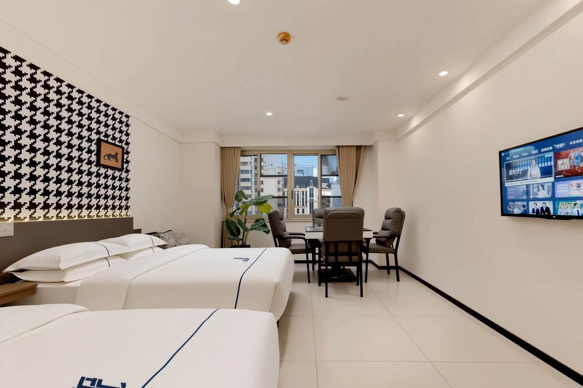 A hotel room with two beds, a dining table with chairs, a TV on the wall, and a large window with a view of buildings.