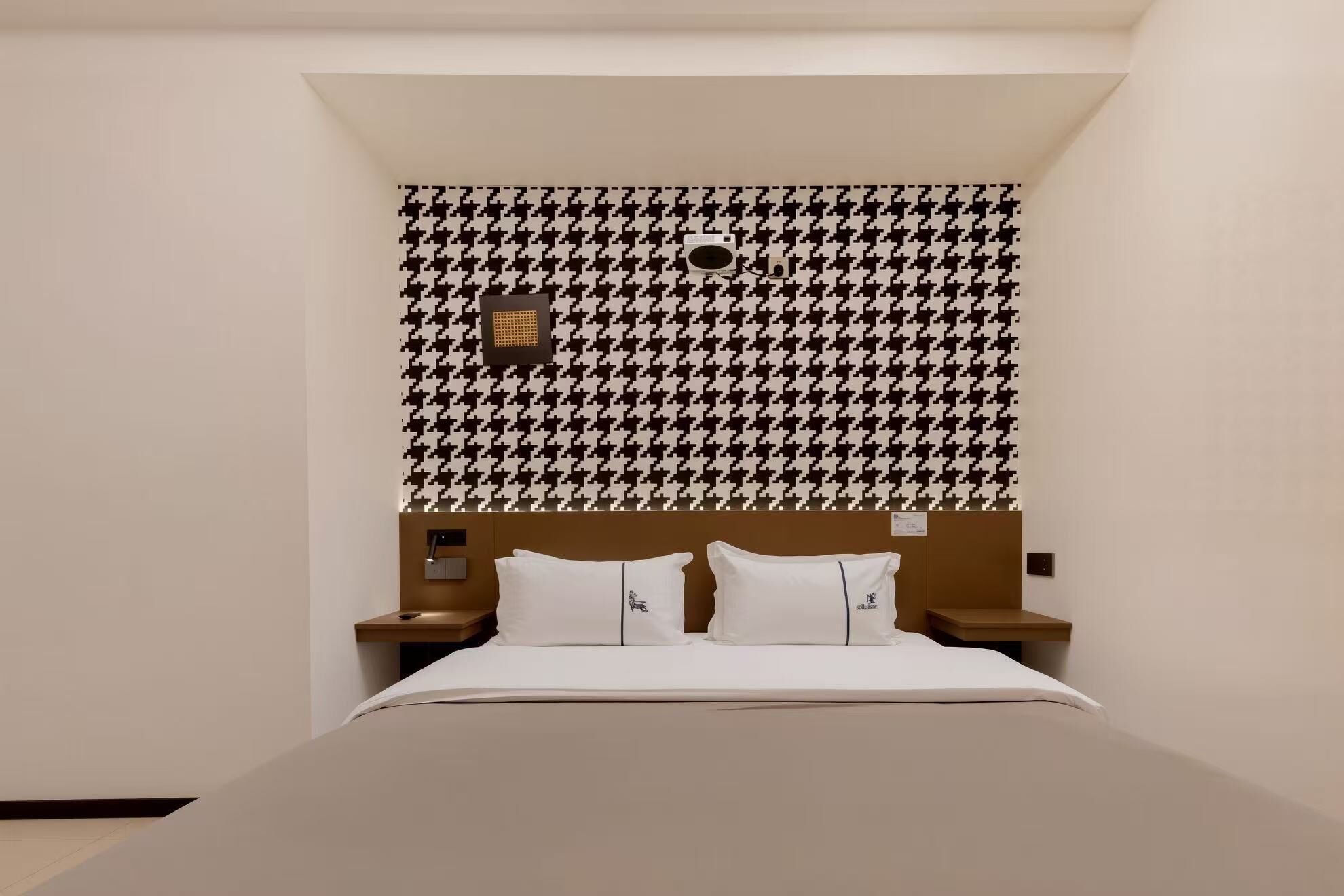 A hotel room with a houndstooth patterned wallpaper, a bed with white bedding, and a wooden headboard.