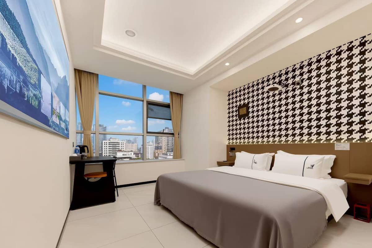 Ein Hotelzimmer mit großem Fenster, einem Bett, einem Schreibtisch und Blick auf die Stadt.