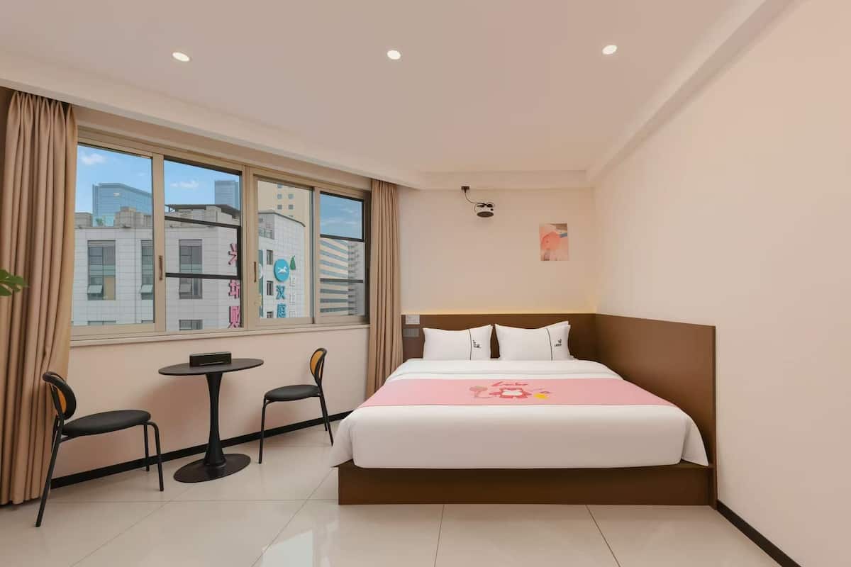 Ein Hotelzimmer mit einem Bett, einem kleinen Tisch, zwei Stühlen, einem Fenster mit Blick auf Gebäude und einer Deckenleuchte.