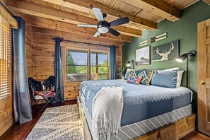 Un dormitorio de cabaña de madera con una cama, un ventilador de techo, una silla y un ventanal con cortinas.