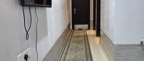 A hallway with a marble-tiled floor, a patterned rug, a mounted TV, and a telephone on a table.