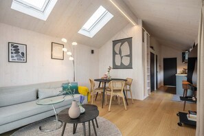 Un salon moderne avec un canapé, une table basse ronde et un coin repas avec des chaises. On peut voir des lucarnes au plafond, des œuvres d’art sur les murs et un coin cuisine à l’arrière-plan.