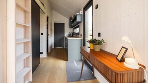 Um corredor estreito com piso de madeira, uma estante embutida, uma área de cozinha com bancada e armários, e uma mesa com luminária e telefone.