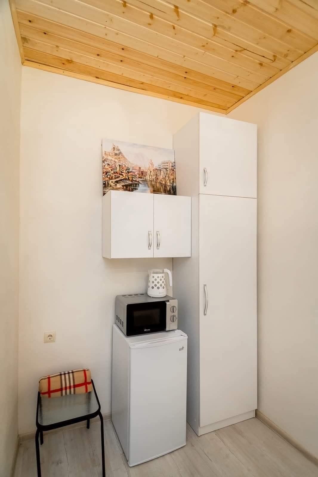 Una habitación compacta con techo de madera, un armario blanco, un refrigerador pequeño blanco, un microondas, una cafetera y una silla.