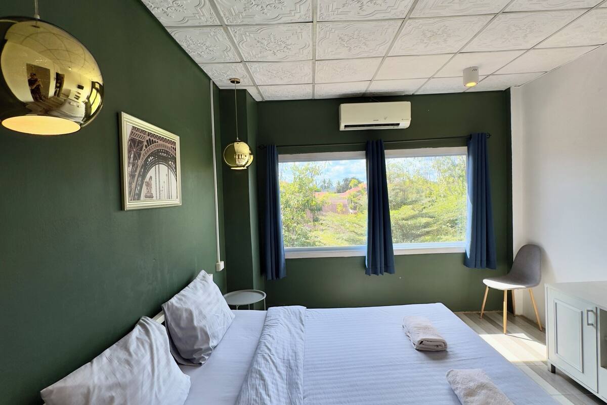 A neatly made bed with pillows, a hanging light fixture, a window with curtains, and a wall-mounted air conditioner.