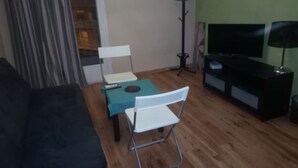A living room with a black sofa, a teal coffee table, a white folding chair, a TV stand with a TV, and a floor lamp. - Cozy 2-bedroom Center in Thessaloniki (Thessaloniki)