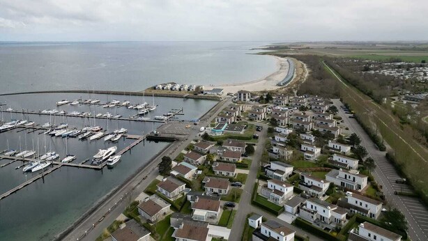 Vista aèria d'una marina costanera amb diverses embarcacions amarrades, una zona residencial amb cases i una platja al fons.