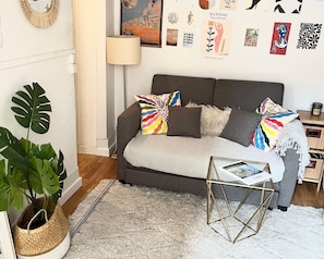 Ein gemütliches Wohnzimmer mit grauem Sofa, bunten Kissen, einem kleinen Beistelltisch, einer Topfpflanze und Wandbildern.