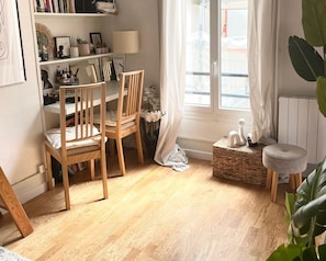 Ein Zimmer mit Holzboden, Schreibtisch mit Stühlen, Bücherregal, Heizkörper und einem Fenster mit Vorhängen.